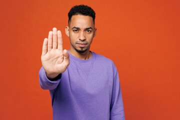 Fototapeta premium Young sad mad strict man of African American ethnicity wear purple sweatshirt casual clothes showing stop gesture with palm isolated on plain red orange background studio portrait. Lifestyle concept.