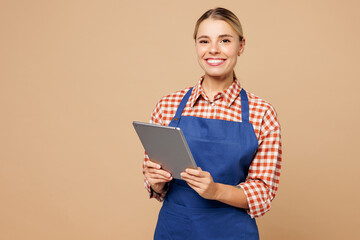 Obraz premium Young woman barista bartender barman employee wears blue apron red shirt work in coffee shop use digital tablet pc computer isolated on plain pastel beige background. Small business startup concept.