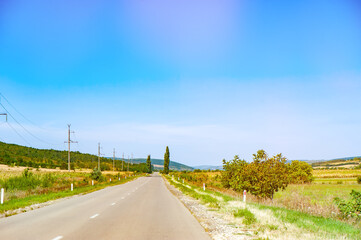 A long, straight rural road with power lines running alongside it, surrounded by fields and trees,...