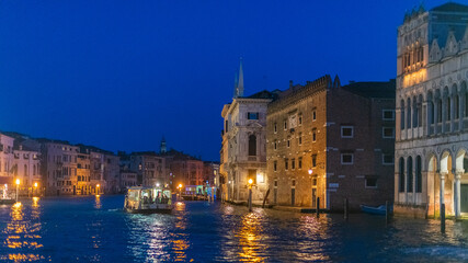 Vue de Venise de nuit depuis le Grand canal avec des quais éclairés et des bâtiments anciens, et le vaporetto qui navigue.