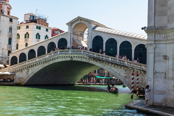 Vue du pont du Rialto à Venise depuis le grand canal avec une gondole qui passe dessous et des touristes sur le pont par une journée ensoleillée.