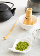 Matcha green tea powder with bamboo spoon and whisk in ceramic bowl with iron kettle on white background.