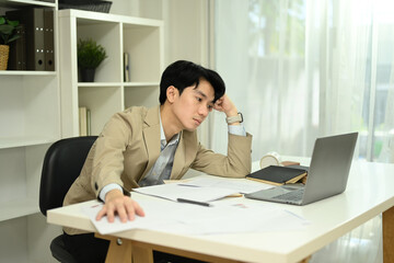 Fototapeta premium Bored asian male employee working inside modern office with laptop and documents