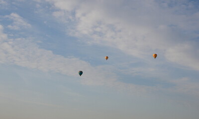 A close up on three colorful balloons flying in the cloudy sky next to some puffy clouds and a small village seen around sunrise on a Polish countryside in summer