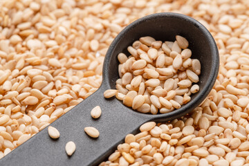 White sesame seeds in black spoon over sesame seeds pile.