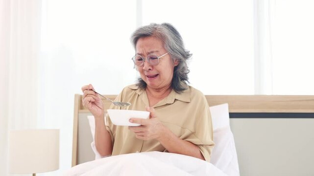 Elderly asian woman tries taste breakfast with dissatisfied expression. She looks at the food in the bowl and feels bored with eating the food in the bowl that is bland lacks flavor and is monotonous.