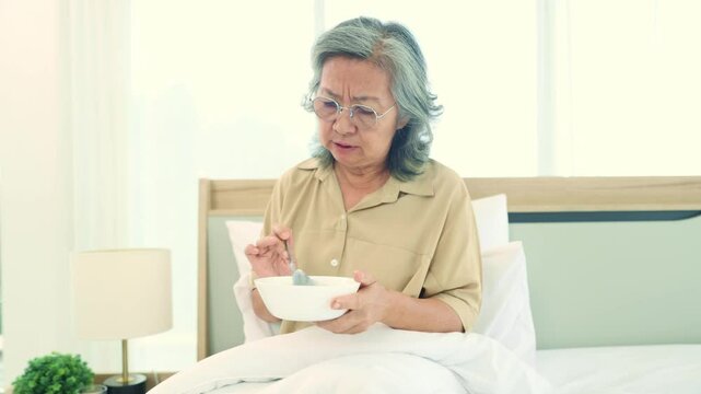 Elderly asian woman tries taste breakfast with dissatisfied expression. She looks at the food in the bowl and feels bored with eating the food in the bowl that is bland lacks flavor and is monotonous.