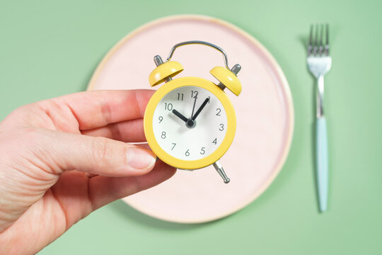 Female hand holding alarm clock against empty plate, intermittent fasting concept.