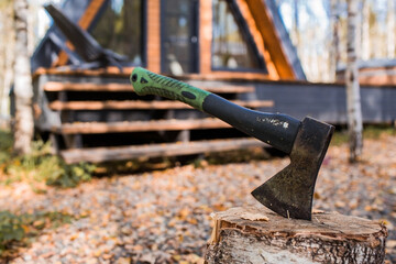 An axe will be stuck into a wooden log. Harvesting firewood for the winter season. Logging on the background of a country house. Lumber, wood-cutting tools.
