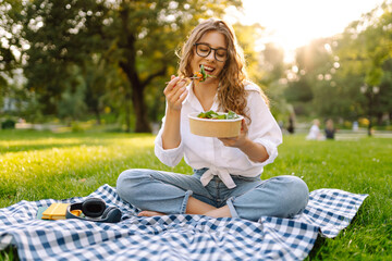 Woman enjoying a poke bowl on a sunny day in a park while sitting on a picnic blanket surrounded by...