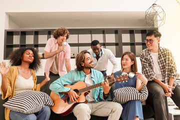 A young, handsome man is happily playing acoustic guitar on a sofa in a domestic house, surrounded by multiracial friends who are singing a song together and sharing a warm moment of togetherness