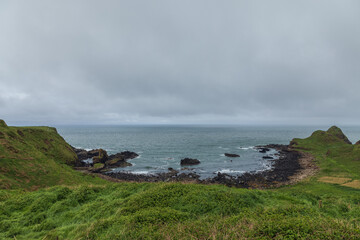 Green hills and rugged rocks surround the Bay of the Cow near Giant's Causeway, meeting the calm Atlantic under grey, cloudy skies