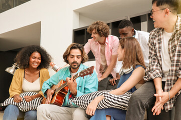 A young, handsome man is happily playing acoustic guitar on a sofa in a domestic house, surrounded by multiracial friends who are singing a song together and sharing a warm moment of togetherness