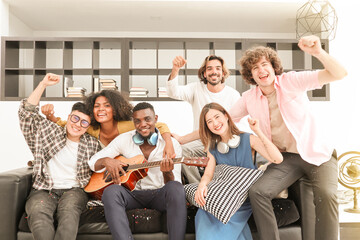 A group of multiracial teenagers playing guitar and singing a song together at the afternoon party. A group of Millennials sitting and raising their hands to celebrate the holiday weekend together.