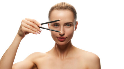 Fototapeta premium Portrait of young beautiful woman posing with two makeup pencil for eyes and lips isolated on white studio background. Concept of natural female beauty, skin care, cosmetics and cosmetology