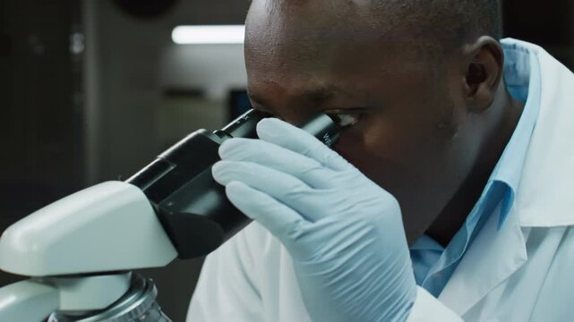 Tilt down shot of African American forensic scientist in gloves and white coat turning microscope lenses while examining blood specimen in crime lab