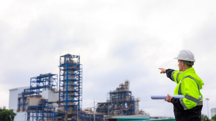 engineer in uniform and hardhat standing pointing finger and holding blueprint at oil refinery plant construction site and sunlight clouds sky backgrounds.