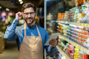Male store employee smiling confidently while holding tablet in grocery aisle. Worker wearing apron...