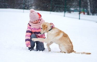 Little Girl And A Golden Retriever Puppy Playing In The Snow Outdoors In Winter, With The Dog Giving A Paw