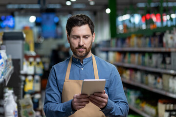 Grocery store worker in apron using digital tablet to manage inventory. Employee stands in...