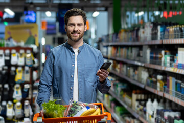 Man in supermarket using phone while shopping, holding basket filled with fresh produce. Brightly...