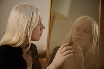 Medium closeup of mysterious young Caucasian woman with albinism looking in mirror and touching her reflection