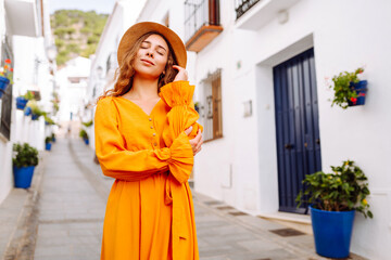 A young traveler woman in a vibrant orange dress and hat enjoys a sunny stroll through a charming white-washed village with flower pots. Concept of lifestyle, vacation, fashion, travel