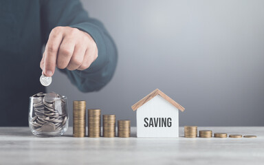 Man drops coins into a glass jar, with stacks of coins and a wooden house model beside him....