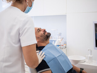 A dental professional in a white uniform assists a patient sitting in a modern clinic. The patient...