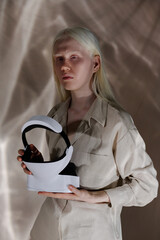 Vertical studio portrait of young Caucasian woman with albinism wearing beige clothes holding VR...