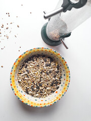 A top view of a colorful bowl filled with various bird seeds next to an empty bird feeder on a white surface.