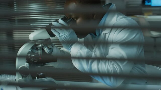 Tilt down shot through window blinds of Black male scientist performing microscopic examination and using digital tablet in forensic laboratory