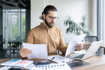 Fototapeta premium Confident male employee analyzing paperwork in contemporary office setting. Professional businessman using laptop for efficient work process. Serious focus on details and strategic planning