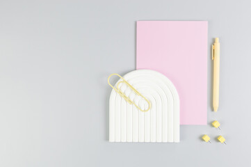 Top view of pink notebook, yellow pen, paperclip, pins, white arch tray on grey background. School, office wallpaper. Flat lay, copy space.