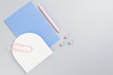 Top view of blue notebook, pink pen, paperclip, pins, white arch tray on grey background. School, office wallpaper. Flat lay, copy space.