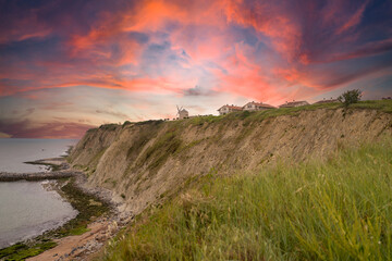 Homes and a windmill on a cliff edge, overlooking the ocean at sunset with a vivid, colorful sky....