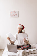 Concerned young man sitting in bed spending time in bedroom at home in Christmas morning. Remote work concept. Guy in Santa hat working with laptop and papers documents