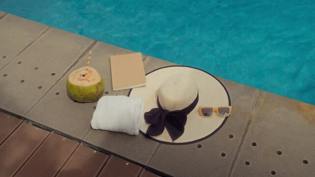 High angle view of straw hat, coconut drink, sunglasses, notebook and white towel left by poolside at resort, no people