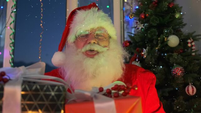 Jolly Santa teasingly sticking out tongue sitting in front of pile of presents next to Christmas tree of shiny ribbons and festive lights. Close up.