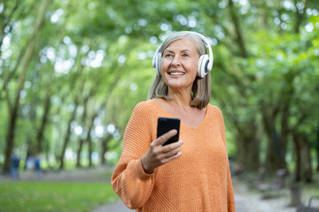 Mature woman listening to music with headphones and smartphone outside in green park. Older female...