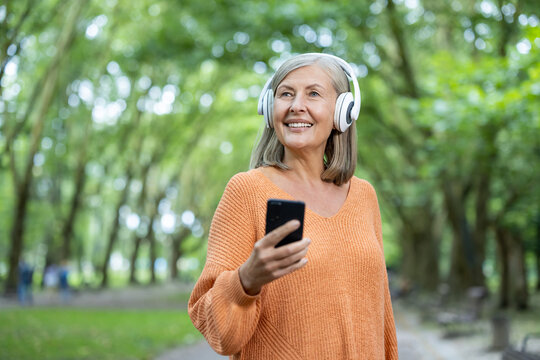 Mature woman listening to music with headphones and smartphone outside in green park. Older female enjoying peaceful moment, smiling and showing joy and relaxation. Perfect for lifestyle concepts.