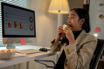 A woman is eating a sandwich while sitting at a desk with a computer monitor in