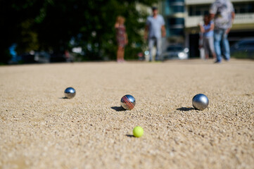 Close-up of petanque balls scattered on gravel with the target ball in focus. The shiny metal balls reflect sunlight, capturing a playful outdoor moment in a park setting