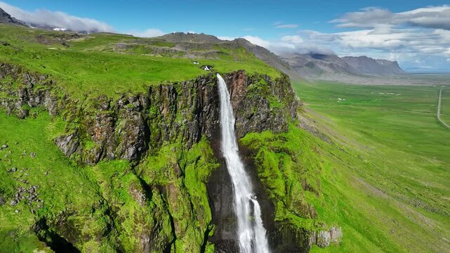 Aerial around shot of Bjarnarfoss waterfall with rainbow, mountains, green meadows and blue sky in the background. Bjarnarfoss waterfall in western Iceland, on sunny day. Summer in Iceland