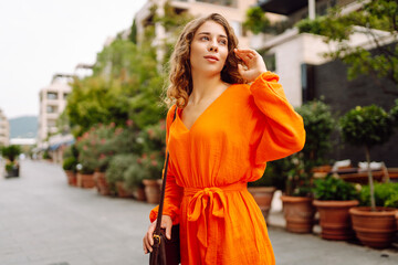 A woman wearing a vibrant orange dress strolls through an elegant city plaza, thoughtfully tucking...