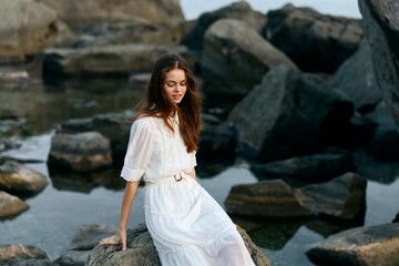 Serene woman in elegant white dress sitting on rugged rock overlooking vast ocean view