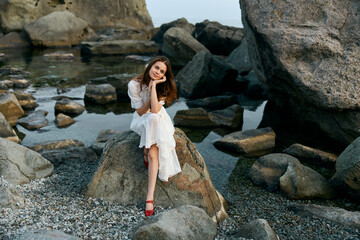 Serene woman in elegant white dress sitting gracefully on a rocky shore with legs crossed beside tranquil waters