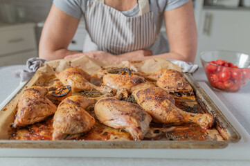Woman with apron serving fresh and homemade cooked roast chicken legs on a baking tray in the kitchen