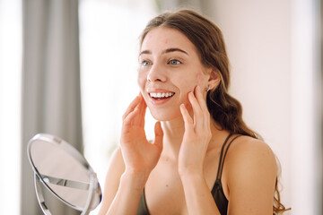 A young woman with problem skin is engaged in her skincare routine, gently touching her face while...