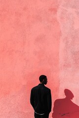 A lone individual stands against a vibrant textured peach wall, casting a shadow and depicting simplicity and contemplation.